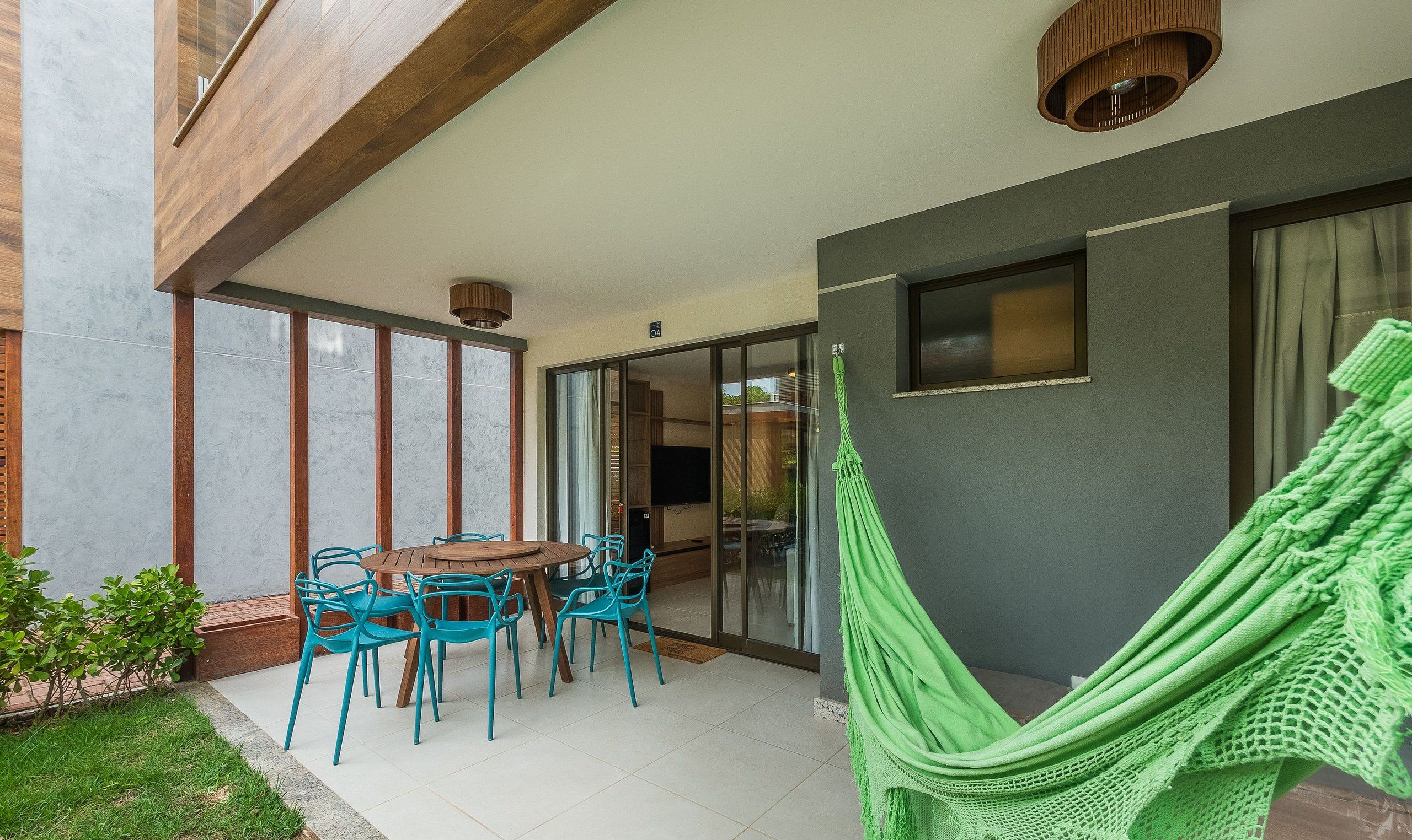 Um pátio com mesa de madeira redonda e cadeiras azuis, uma rede verde e vista do interior através de portas de vidro.