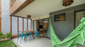 A patio with a round wooden table and blue chairs, a green hammock, and a view of the interior through glass doors.