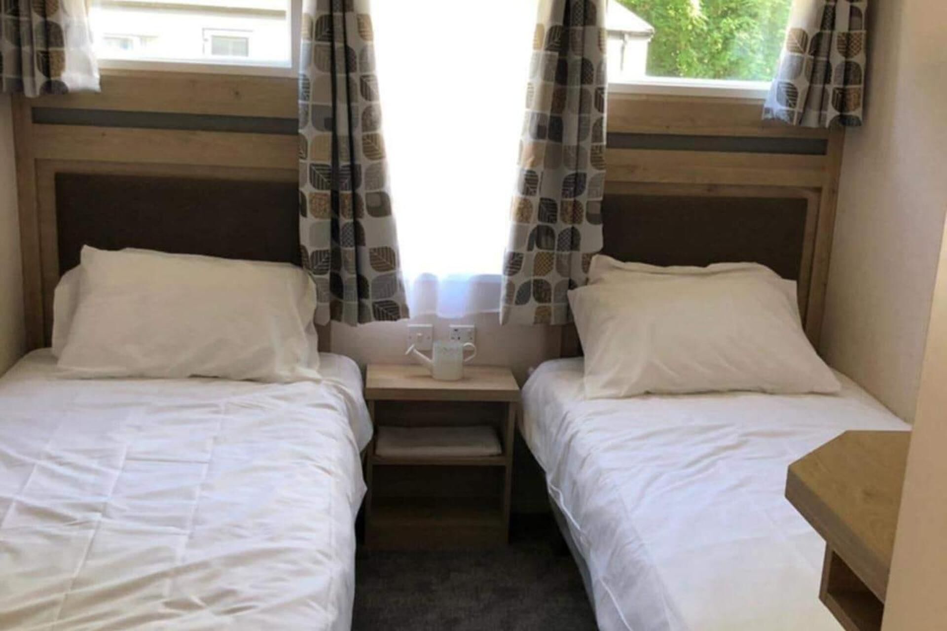 A room with two single beds, a small bedside table with a cup, and curtains with a leaf pattern.