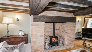 Un salon chaleureux avec des poutres apparentes, une cheminée en briques dotée d’un poêle à bois, un canapé agrémenté de coussins à motifs floraux, une table d’appoint avec une lampe et un meuble pour la télévision.