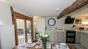 Un coin repas avec une table en bois dressée pour quatre personnes, un vase blanc orné de fleurs et, au fond, une cuisine équipée d’appareils modernes.