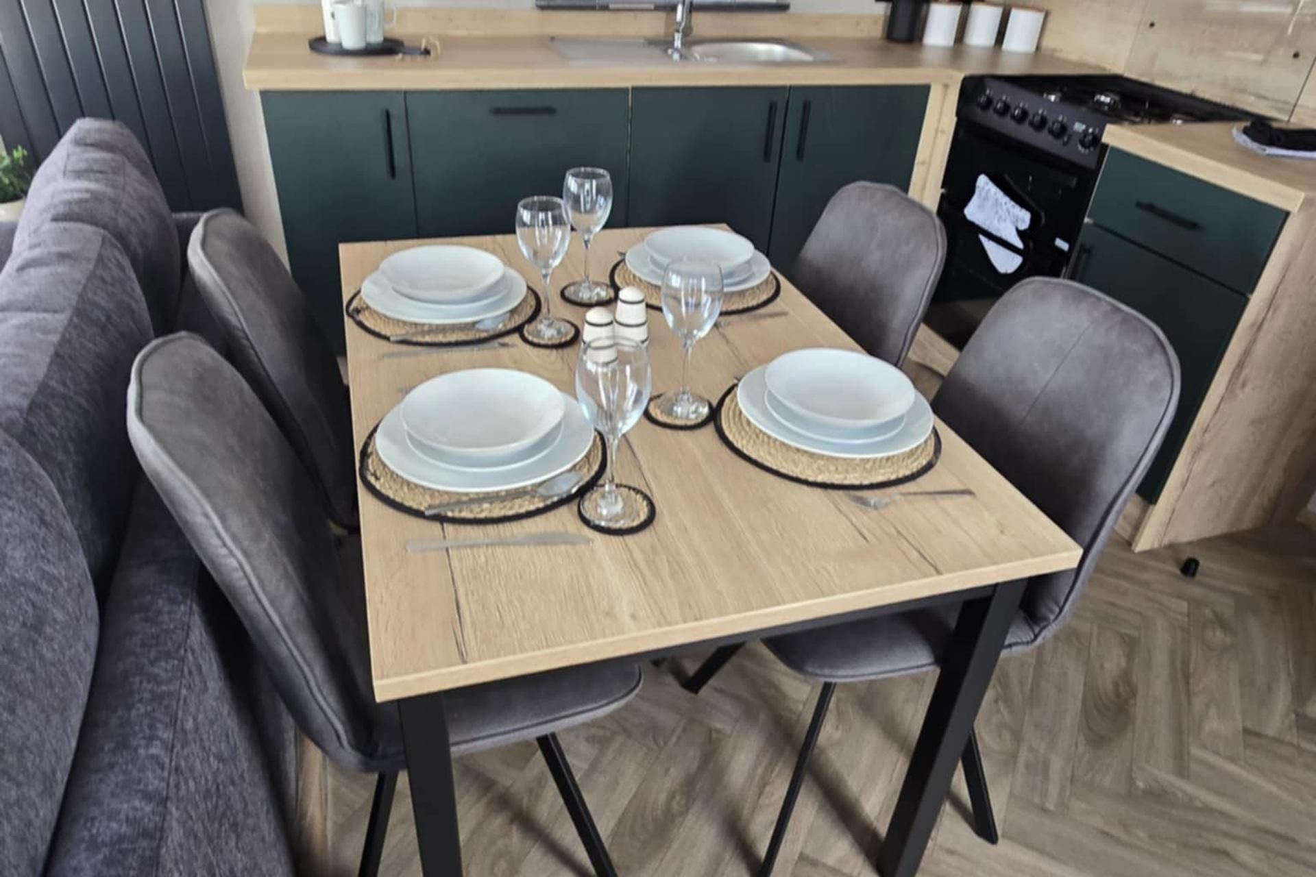 Mesa de comedor dispuesta con platos, vasos y cubiertos en una cocina moderna con armarios oscuros y suelo de madera.