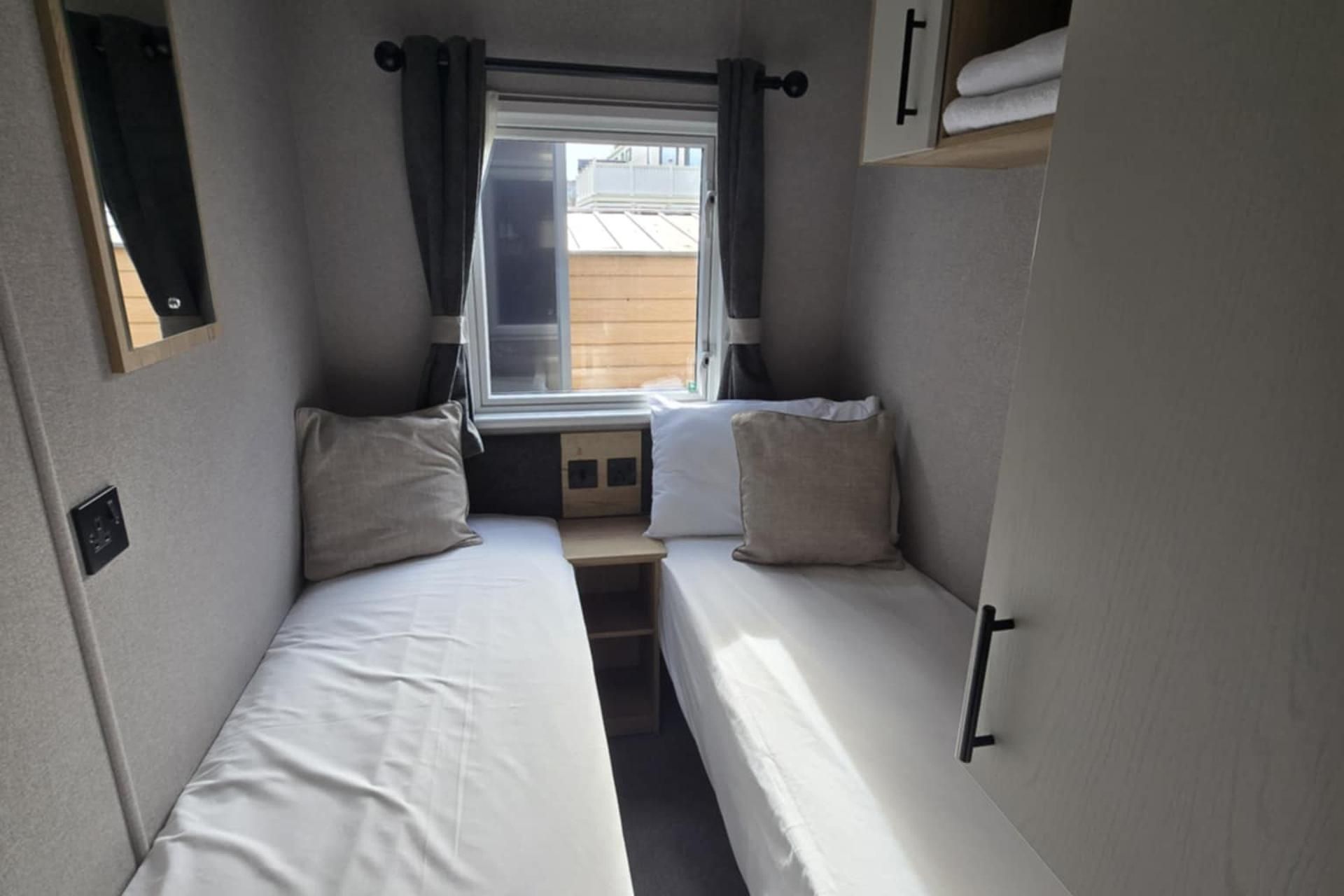Habitación pequeña con dos camas individuales, una ventana con cortinas, una mesita y un armario de pared.