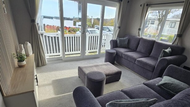 A modern living room with a grey sofa set, a round ottoman, and a wooden sideboard with decorative items. - Dove Close 15 Hoburne Devon Bay (Paignton)