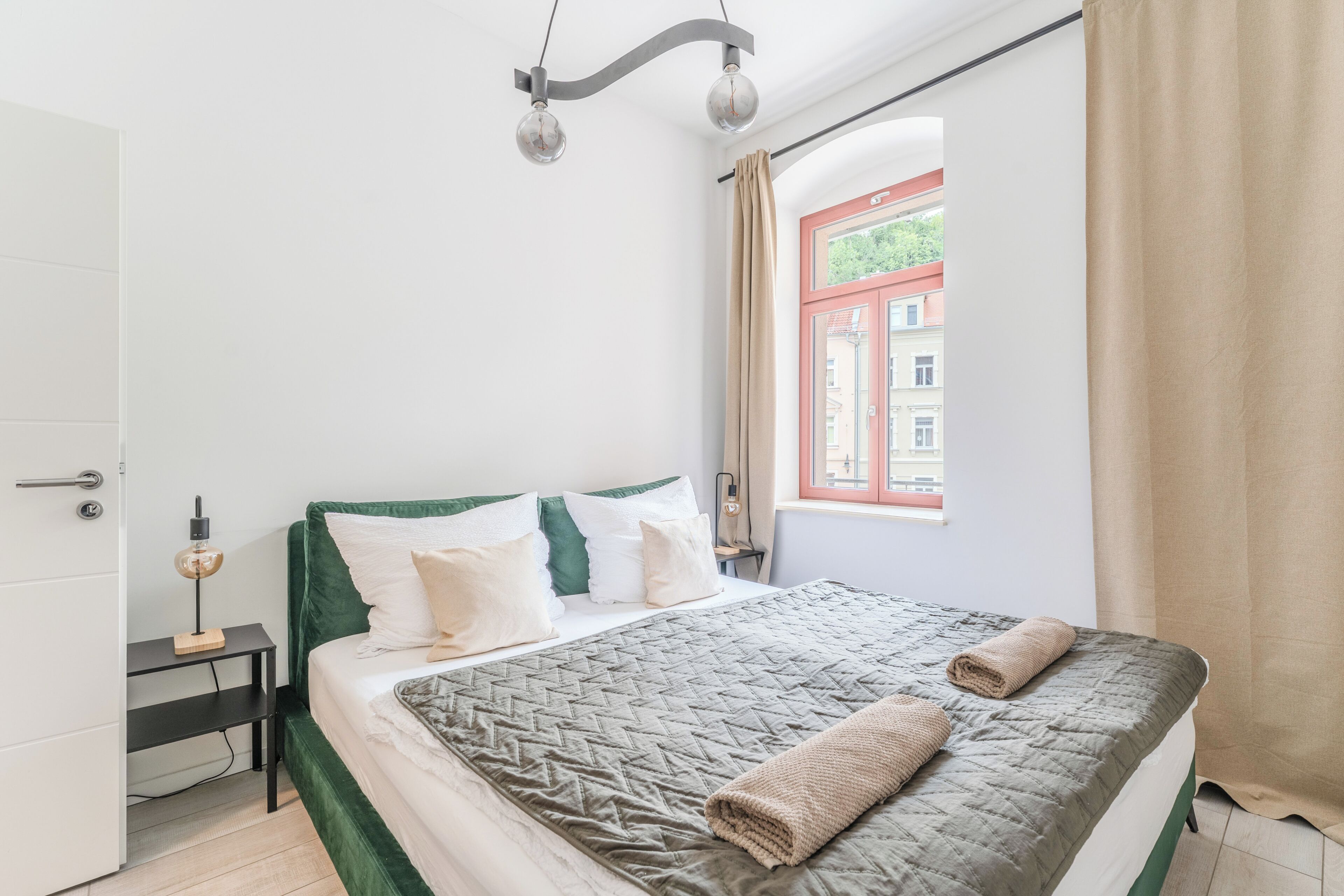 A neatly made bed with a quilted cover and two rolled towels, a bedside table with a lamp, and a window with curtains.