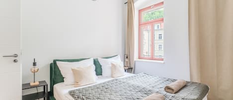 Ein ordentlich bezogenes Bett mit gesteppter Decke und zwei zusammengerollten Handtüchern, ein Nachttisch mit Lampe sowie ein Fenster mit Vorhängen.
