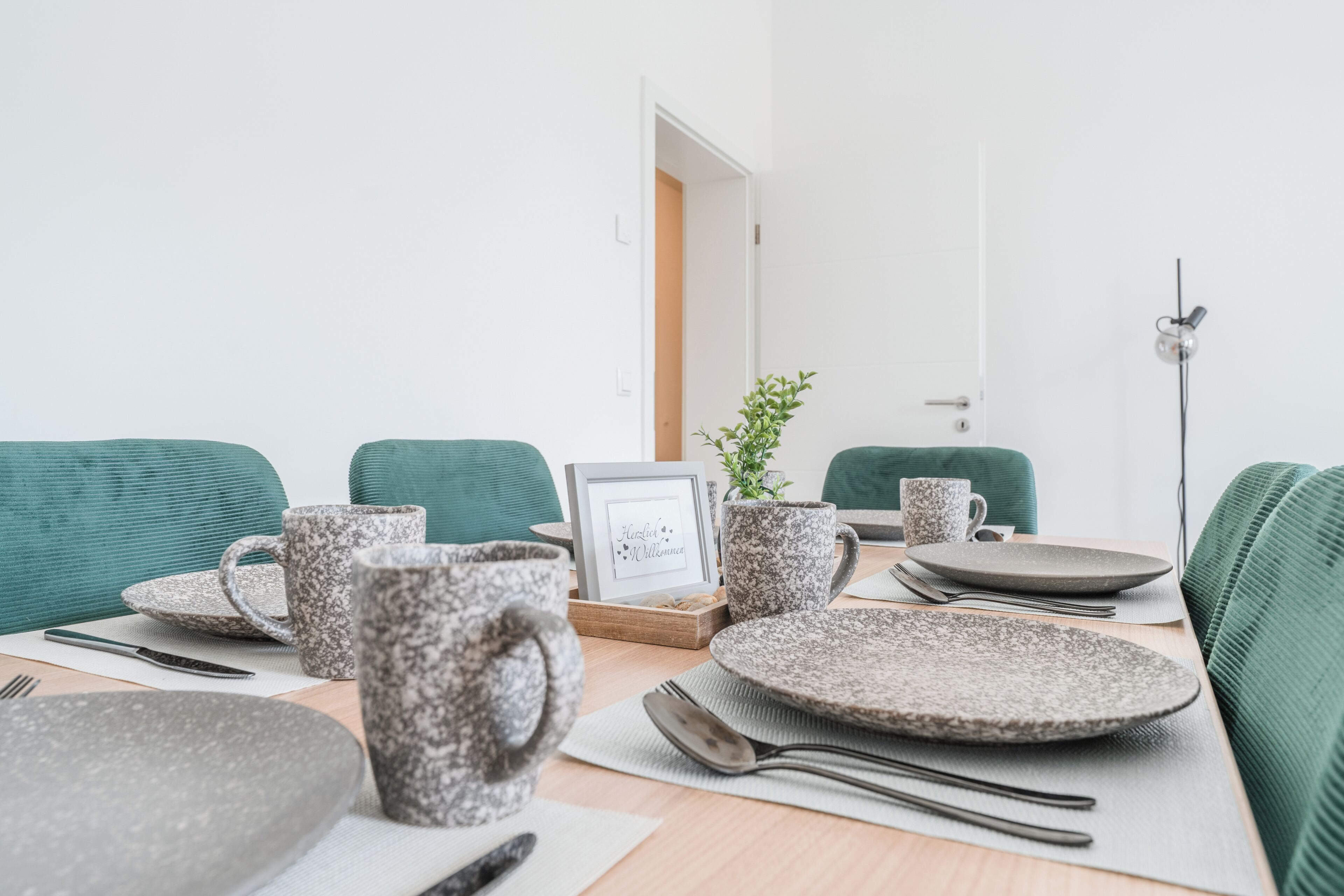 Une table à manger dressée avec des assiettes, des tasses et des couverts, agrémentée d’un tableau encadré et d’une plante en pot.