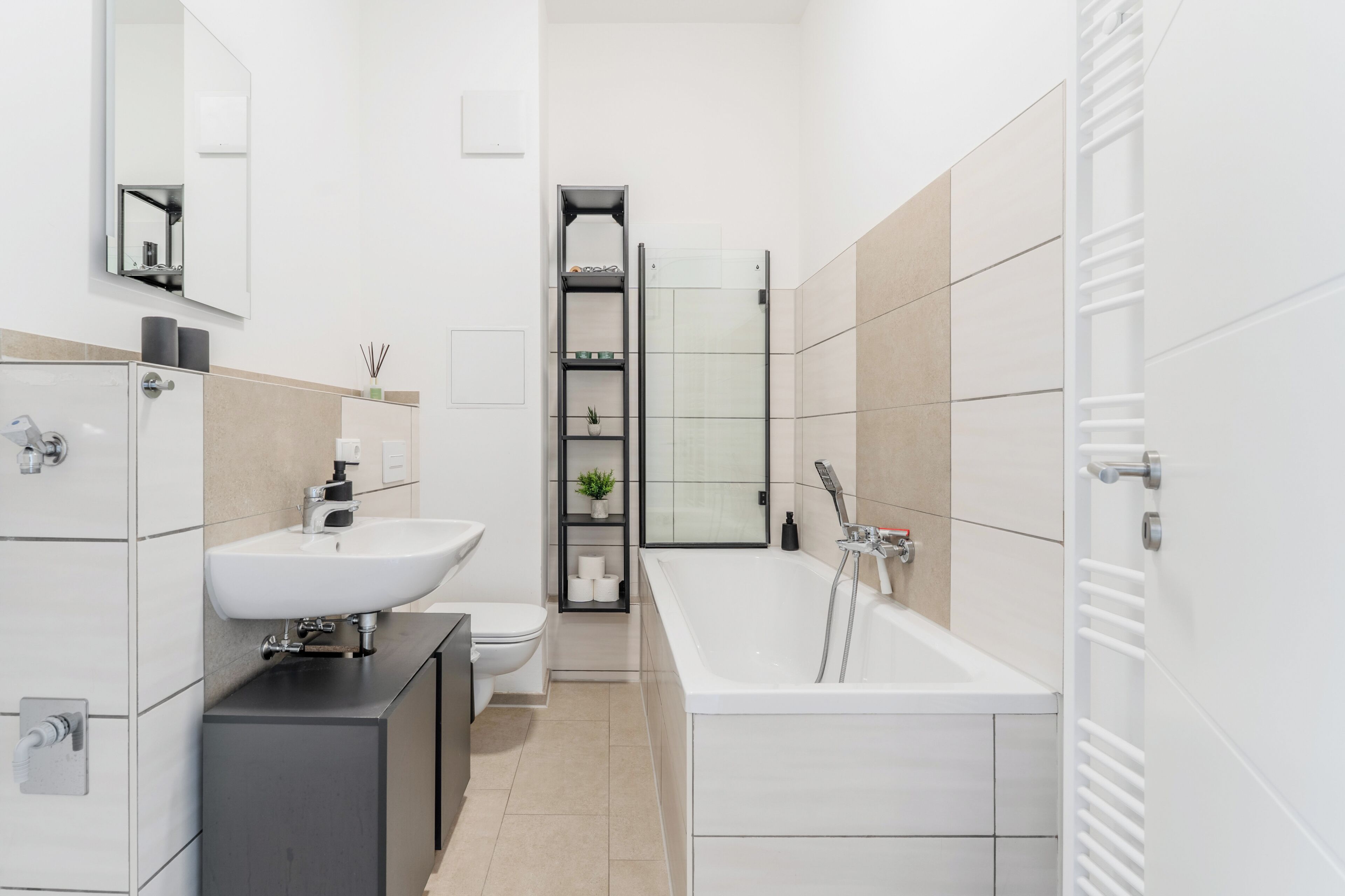 Une salle de bain moderne avec un lavabo, des toilettes et une baignoire, tous de couleur blanche. On y trouve une étagère avec des articles de toilette et une plante en pot.