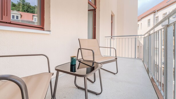 A balcony with two chairs and a small table, a metal railing, and a view of neighboring buildings.