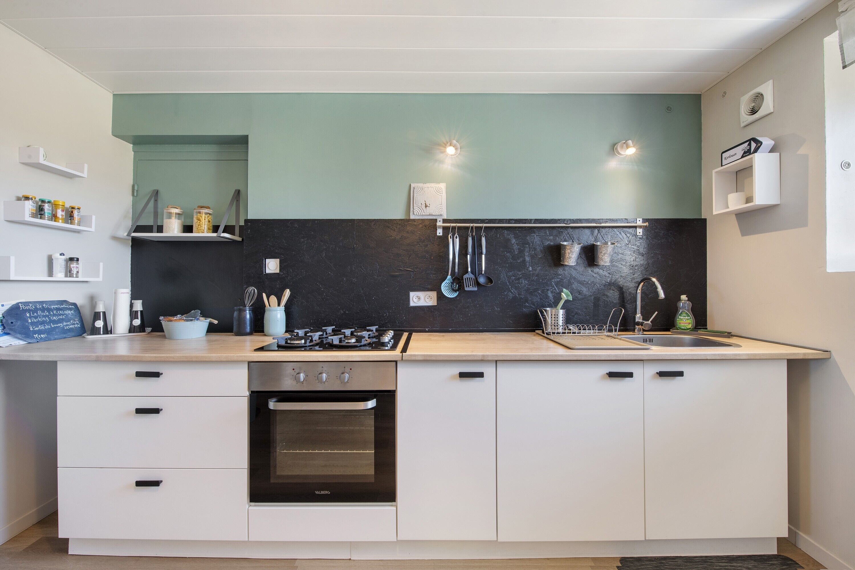 A modern kitchen with a built-in oven, a sink, and a stove. There are cabinets and shelves with kitchen utensils and jars.