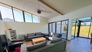 Ein modernes Wohnzimmer mit Sofa, Couchtisch und Fernseher. Große Fenster und ein Deckenventilator sind vorhanden.