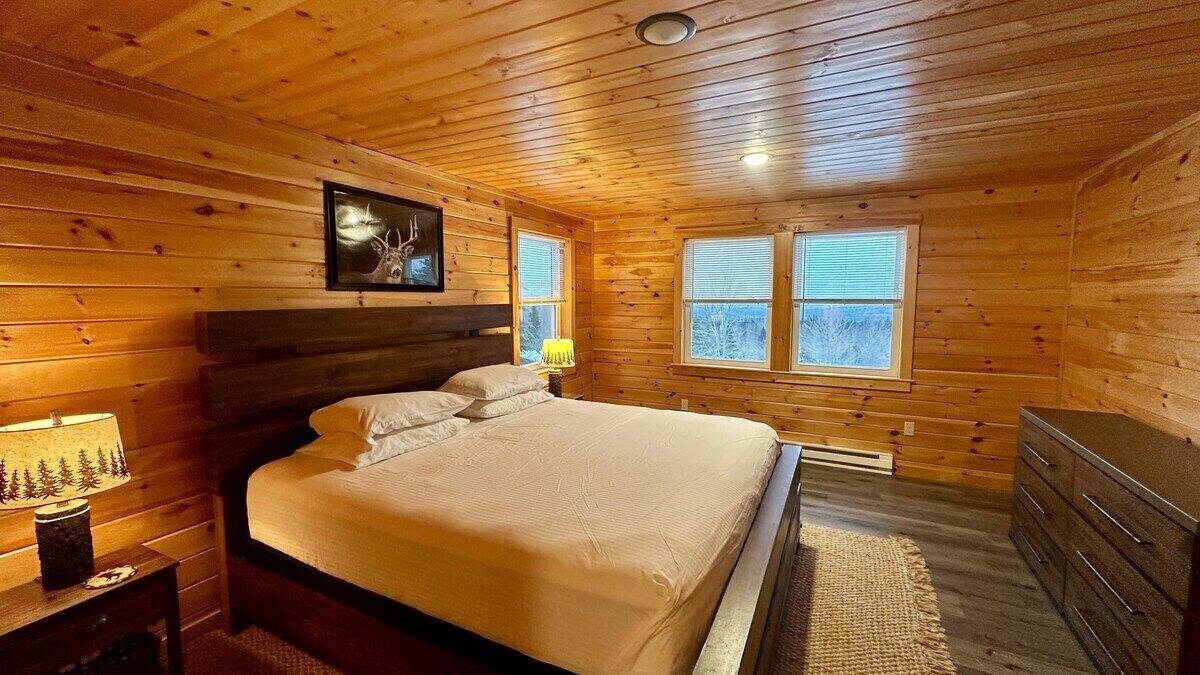 A wooden cabin bedroom with a bed, nightstand, dresser, and windows.