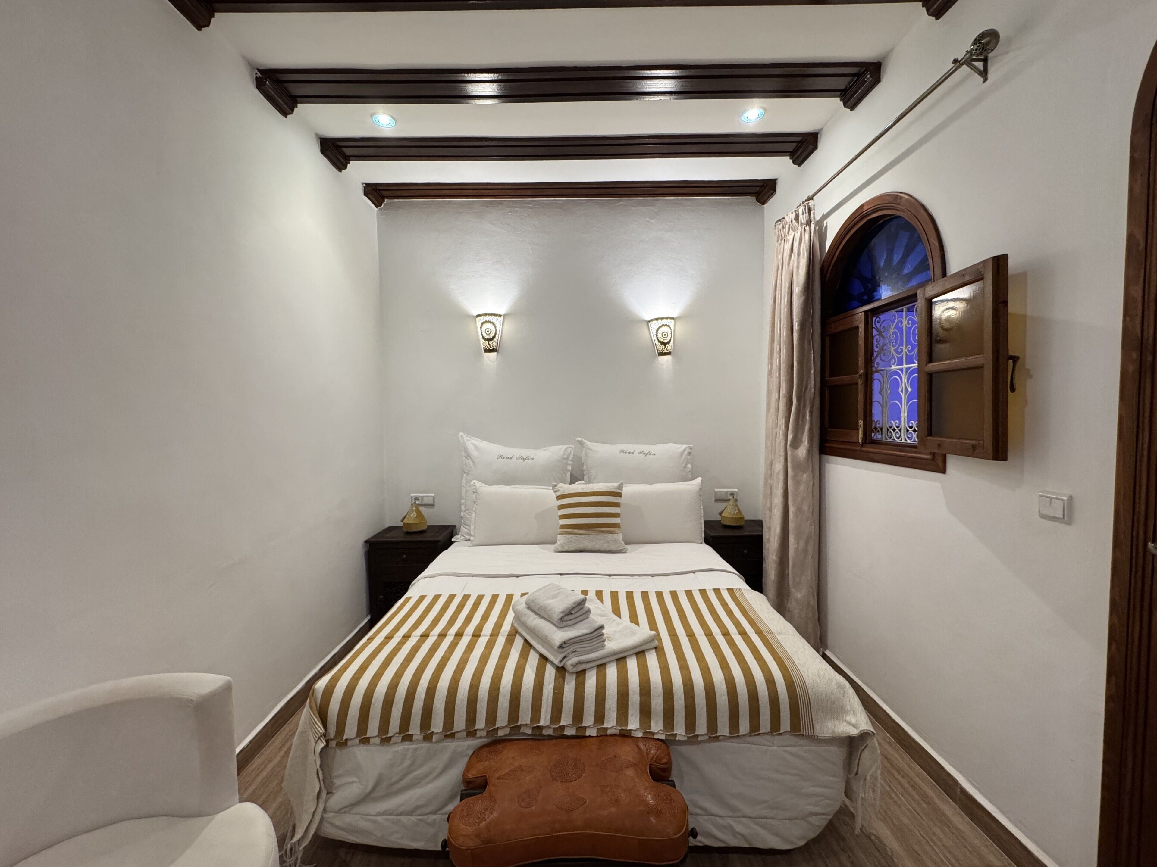 Un dormitorio con una cama, dos mesitas de noche, una lámpara de pared y cortinas en la ventana.