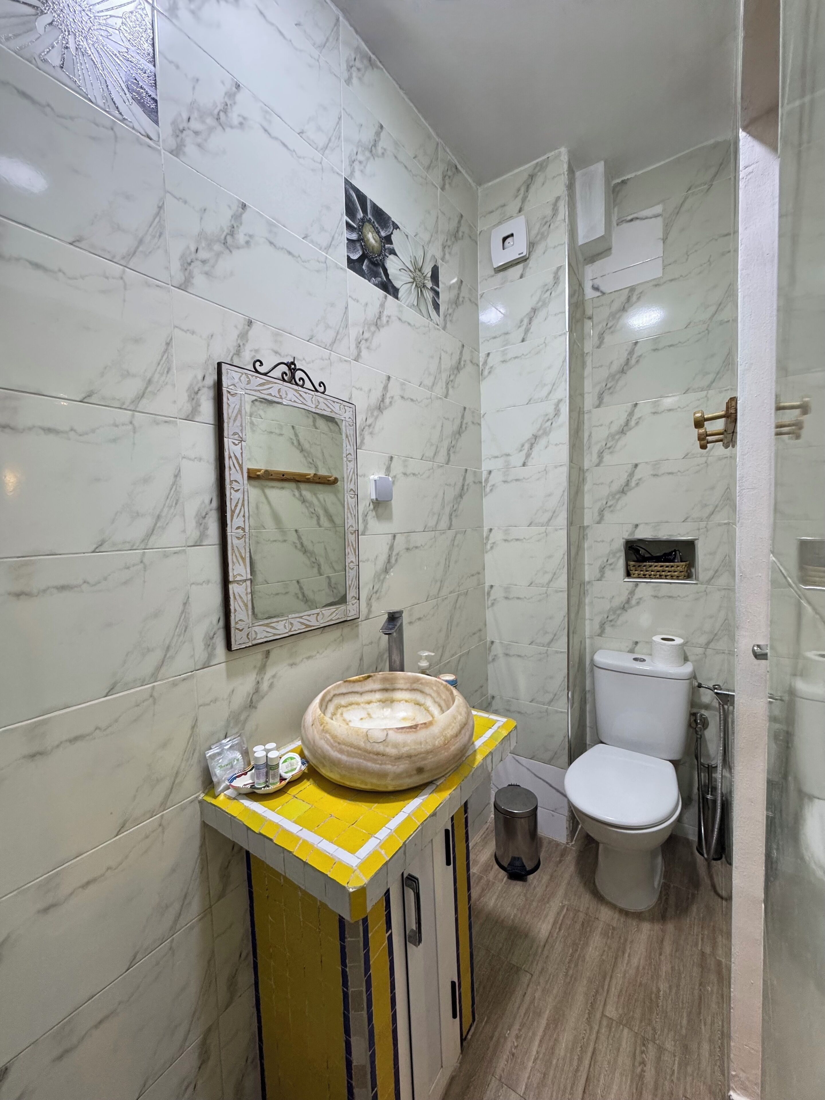 Casa de banho com paredes de mármore, toucador amarelo, um espelho grande, sanita e zona de duche.