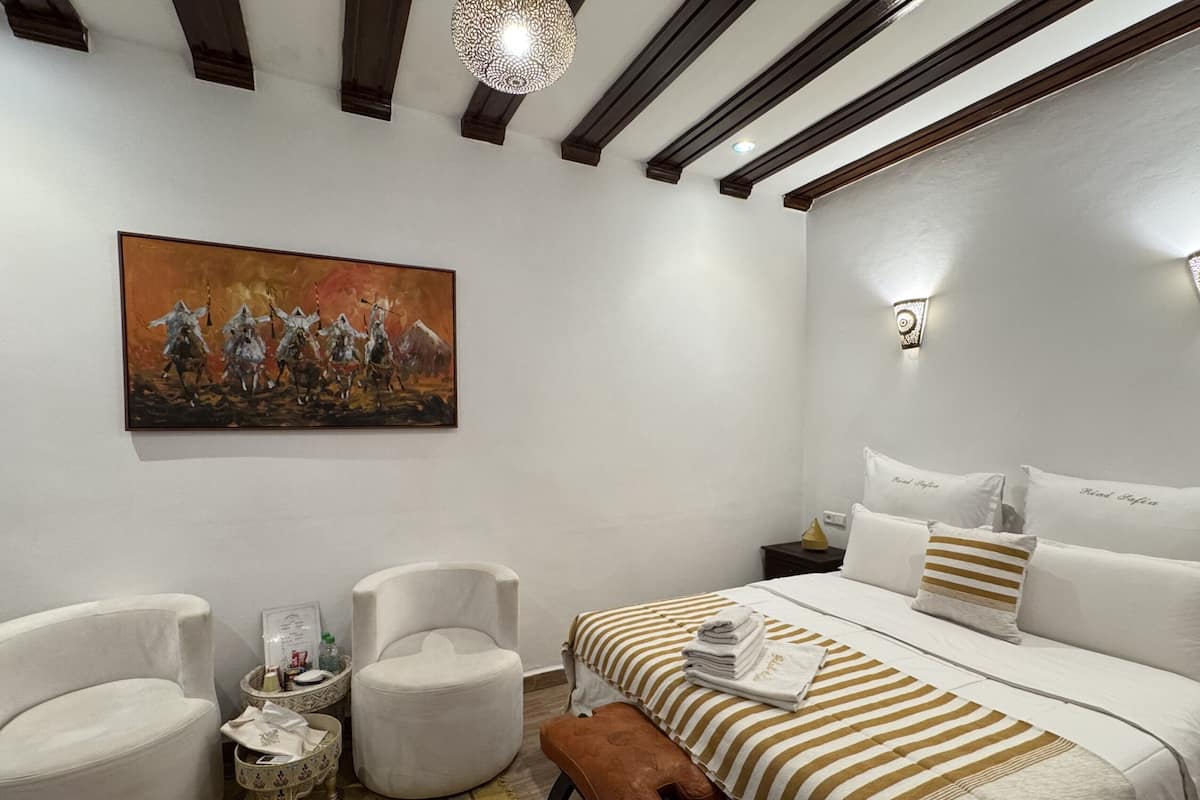 A modern hotel room with a large bed, two armchairs, a small round table, and a painting on the wall.