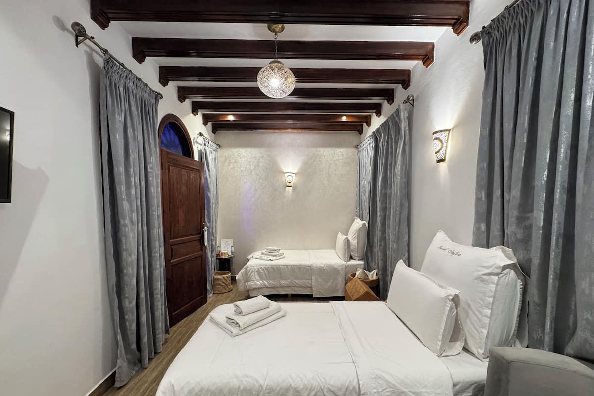 A hotel room with a wooden ceiling, two beds with white linens, grey curtains, a wall-mounted light fixture, and a wooden door.