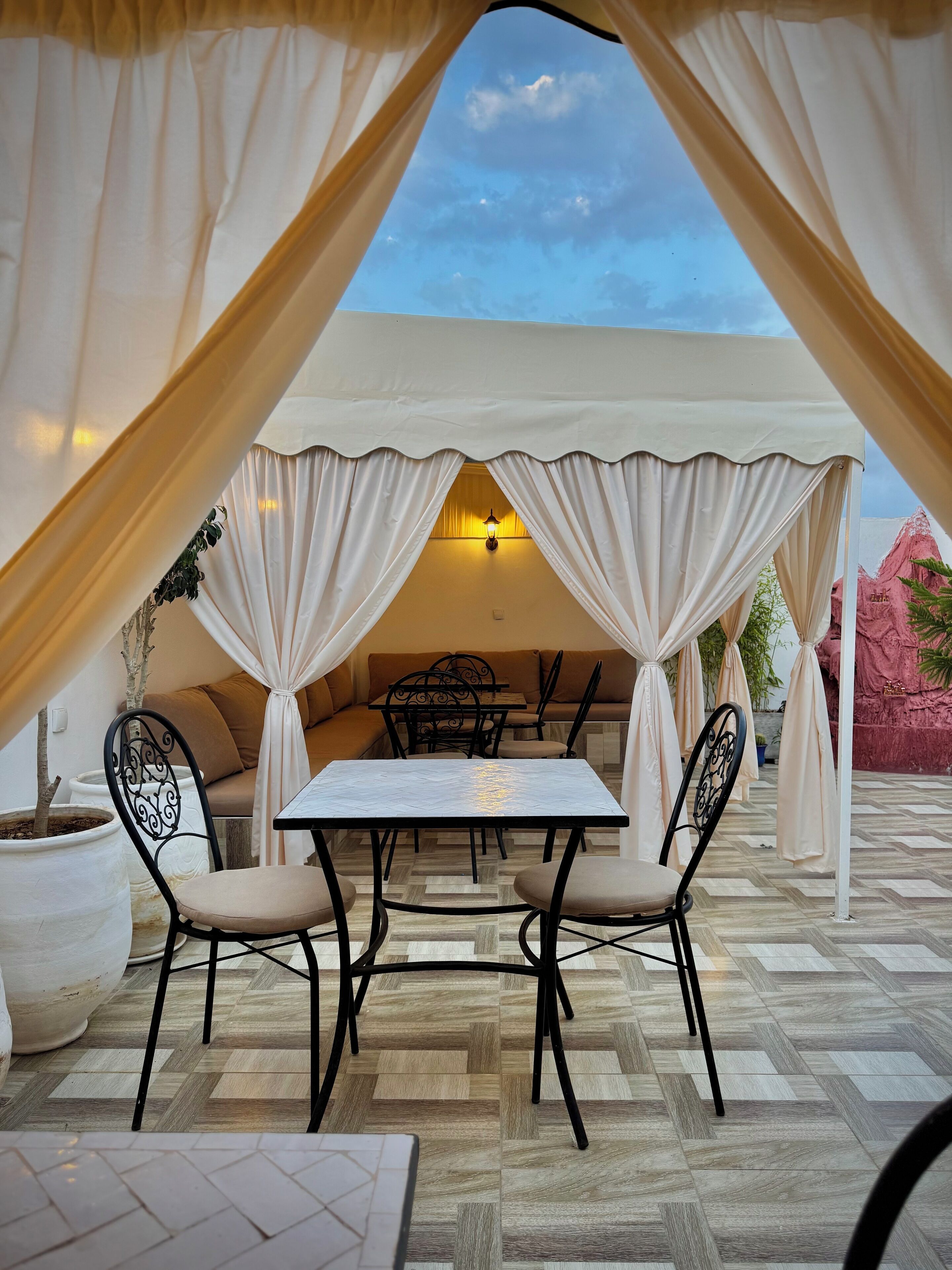 Un área de estar al aire libre con mesa y sillas, decorada con cortinas blancas, bajo un dosel blanco.