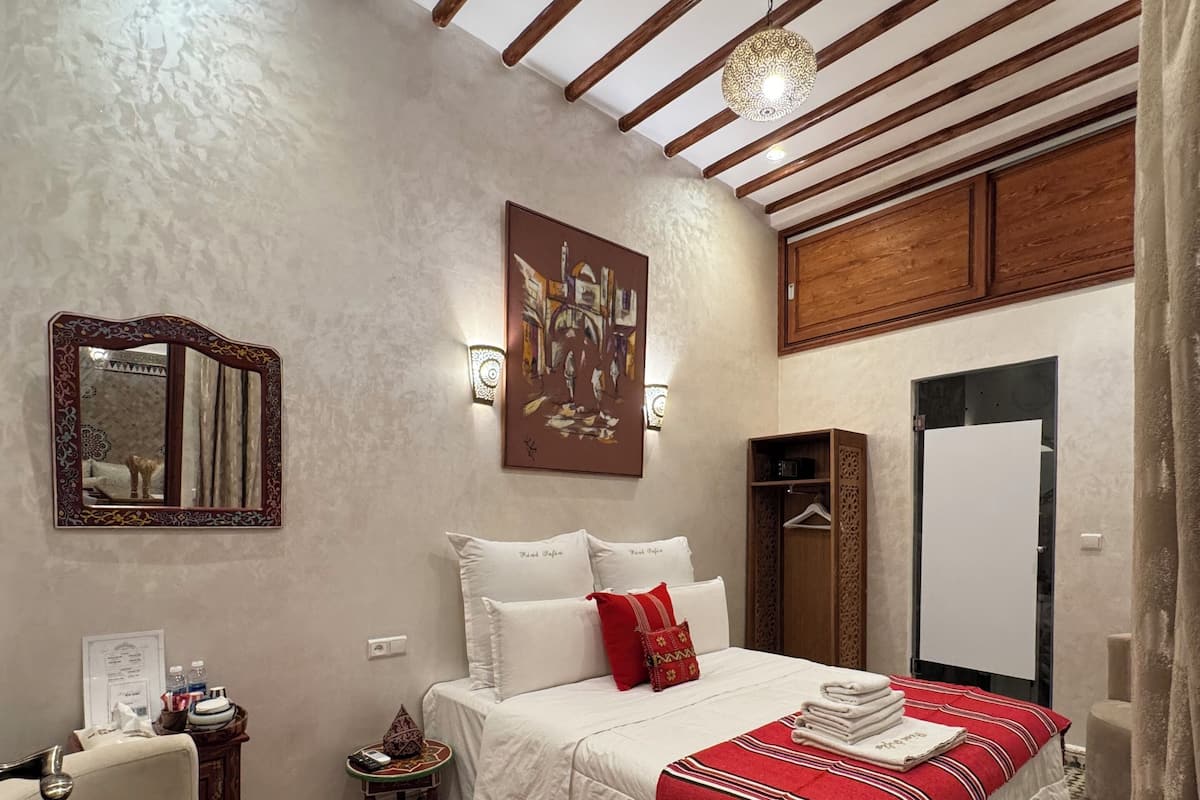 A hotel room with a checkered floor, a bed with white and red bedding, a red throw blanket, a wooden ceiling, and a painting on the wall.