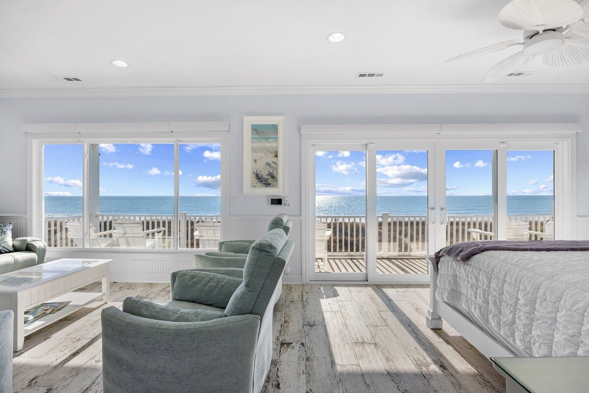Ein Schlafzimmer mit großem Fenster zum Meer, einem Bett, einer Sitzecke mit Stühlen und einem Deckenventilator.