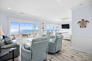 Un salon spacieux avec vue sur l’océan, doté d’un ventilateur de plafond et agrémenté d’une décoration murale représentant des tortues.