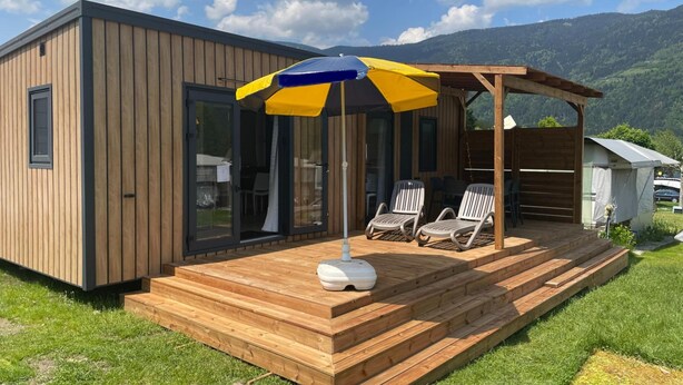 A modern wooden cabin with a deck and outdoor seating area, featuring a large umbrella and a view of the surrounding mountains. - Mobile Home in Austrian Countryside (Ossiach)