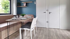 A modern dining area with a wooden table set for four, a bench, and a white chair. There is a window with a view of greenery outside. - Mobile Home in Austrian Countryside (Ossiach)