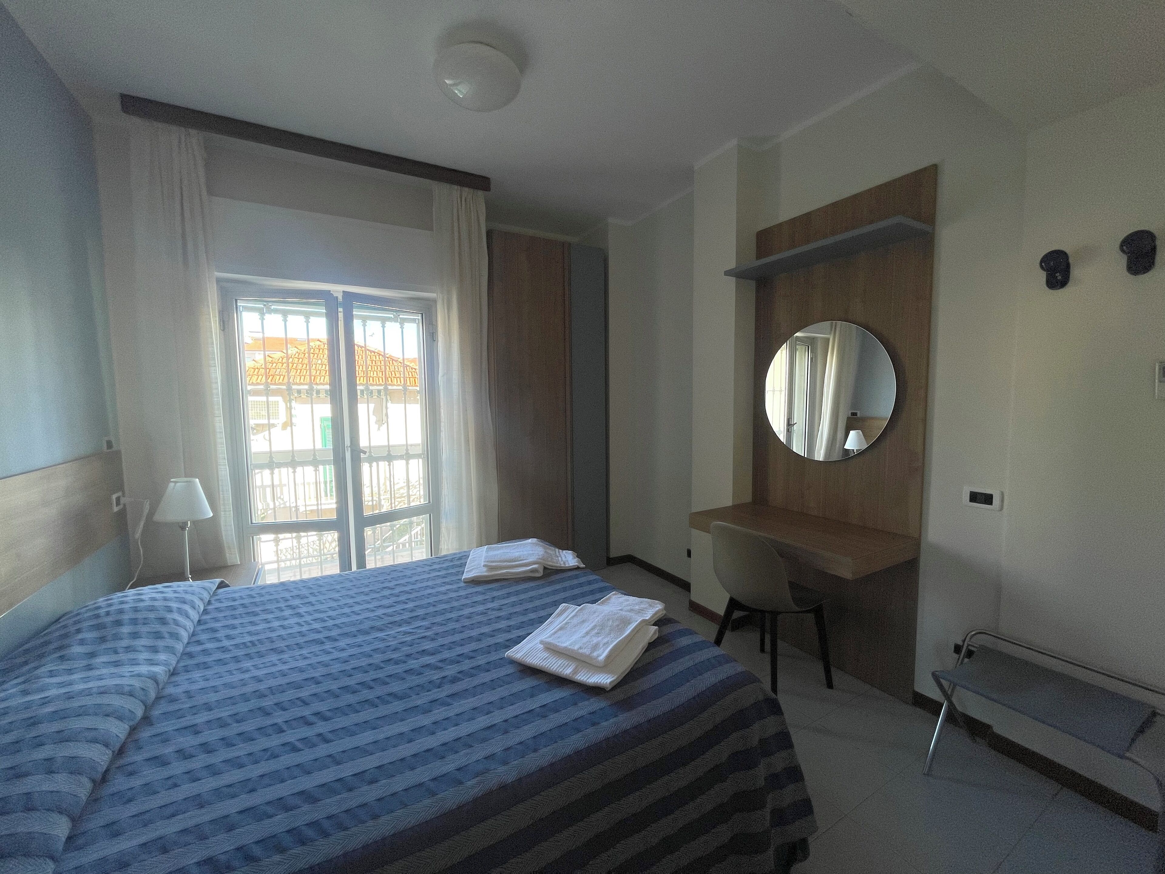 Une chambre d’hôtel avec un lit, un bureau, une chaise, un miroir rond et une fenêtre avec des rideaux.