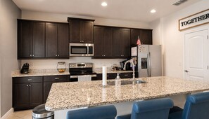 A modern kitchen with dark wood cabinets, stainless steel appliances, granite countertops, and a bar area with blue chairs. - Casa Encore Resort/Piscina privativa/5 Quartos (Kissimmee)