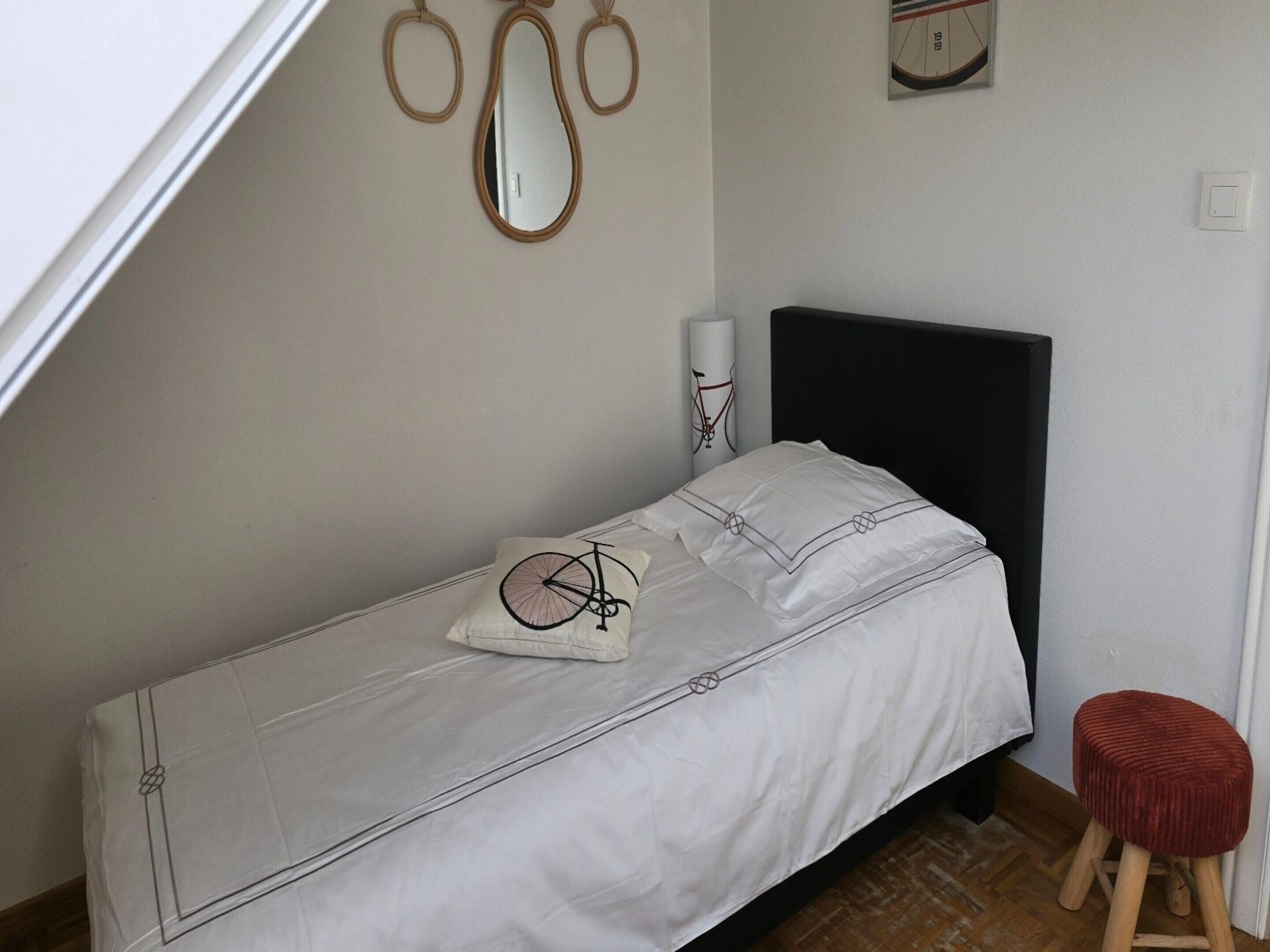 A small, neatly made bed with a white bedspread and a pillow featuring a bicycle design. There is a small stool and a wall-mounted mirror above the bed.