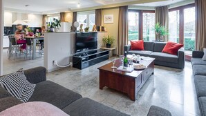 A modern living room with a flat-screen TV, grey sofas, a coffee table, and a dining area with chairs. - Group Accommodation in Borgloon Hof Van Kalenberg (Borgloon)