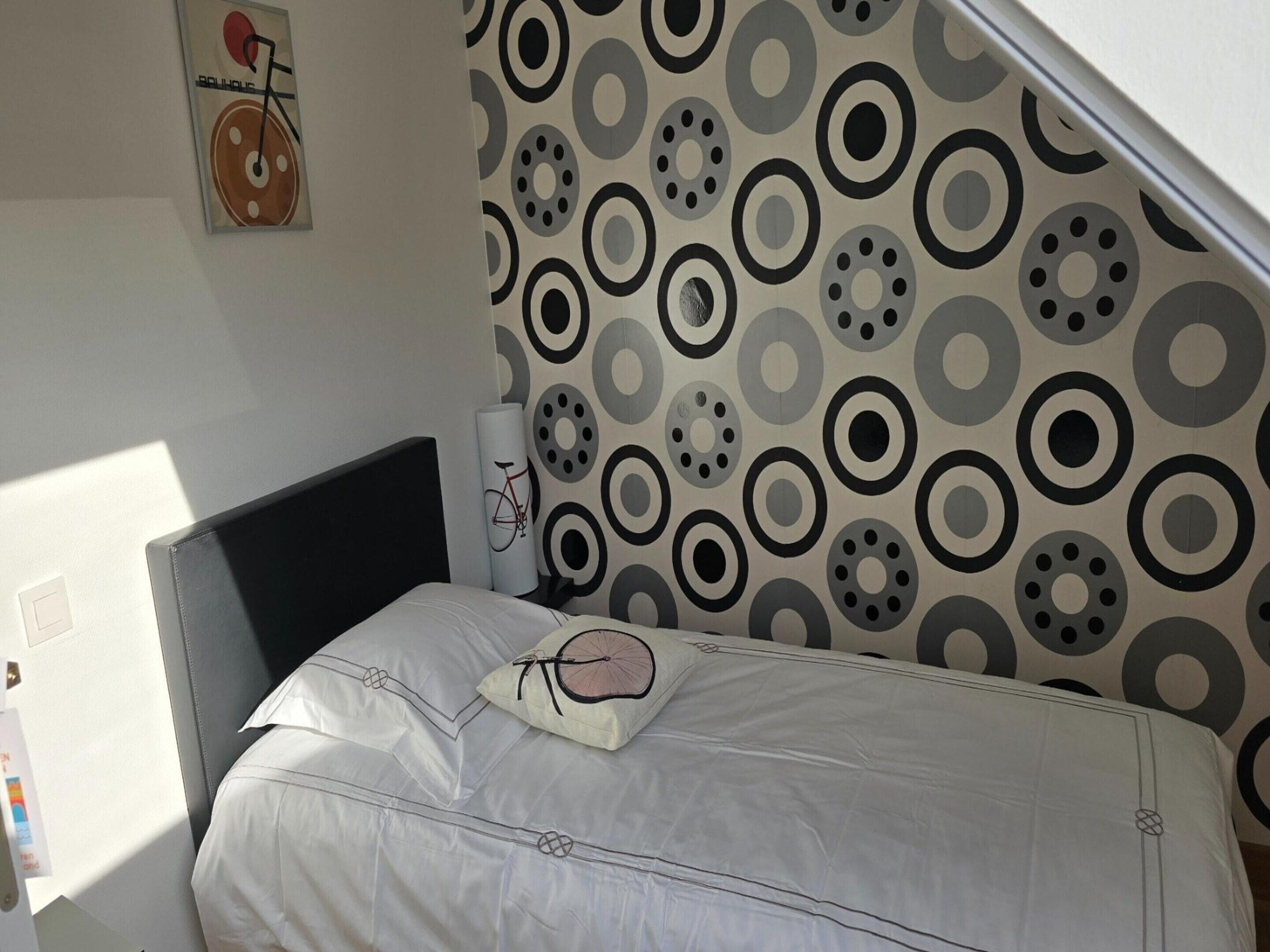 Een slaapkamer met een bed, een kussen met fietsmotief, een wandklok en behang met een patroon.