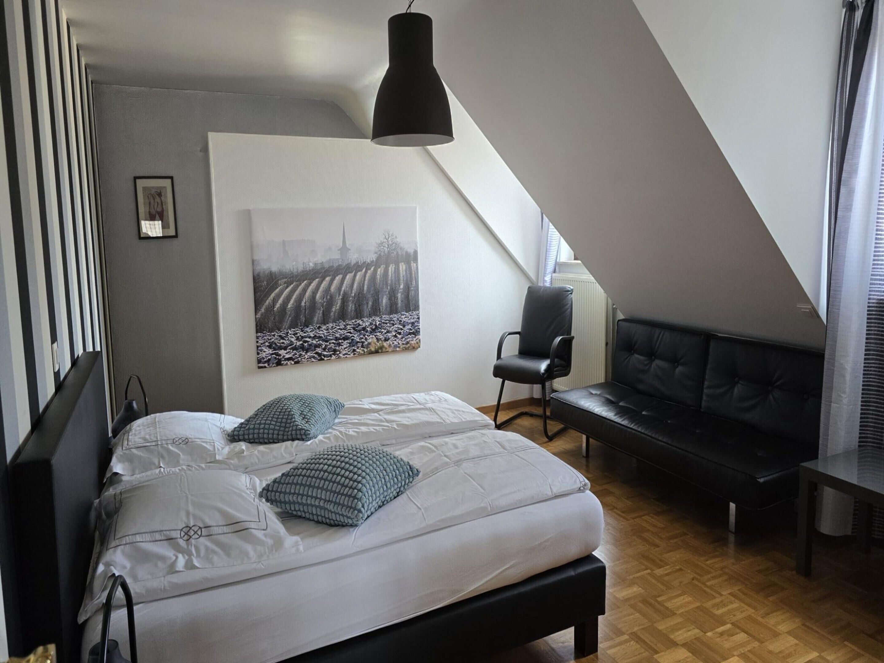 Un dormitorio con una cama, una silla, un sofá, un cuadro y una lámpara colgante.