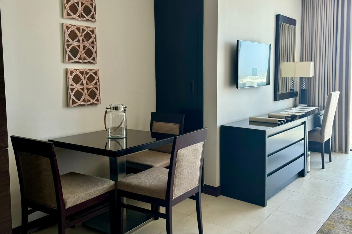 Una habitación de hotel moderna con una mesa de comedor, sillas, un escritorio con silla, un televisor y un ventanal con cortinas.