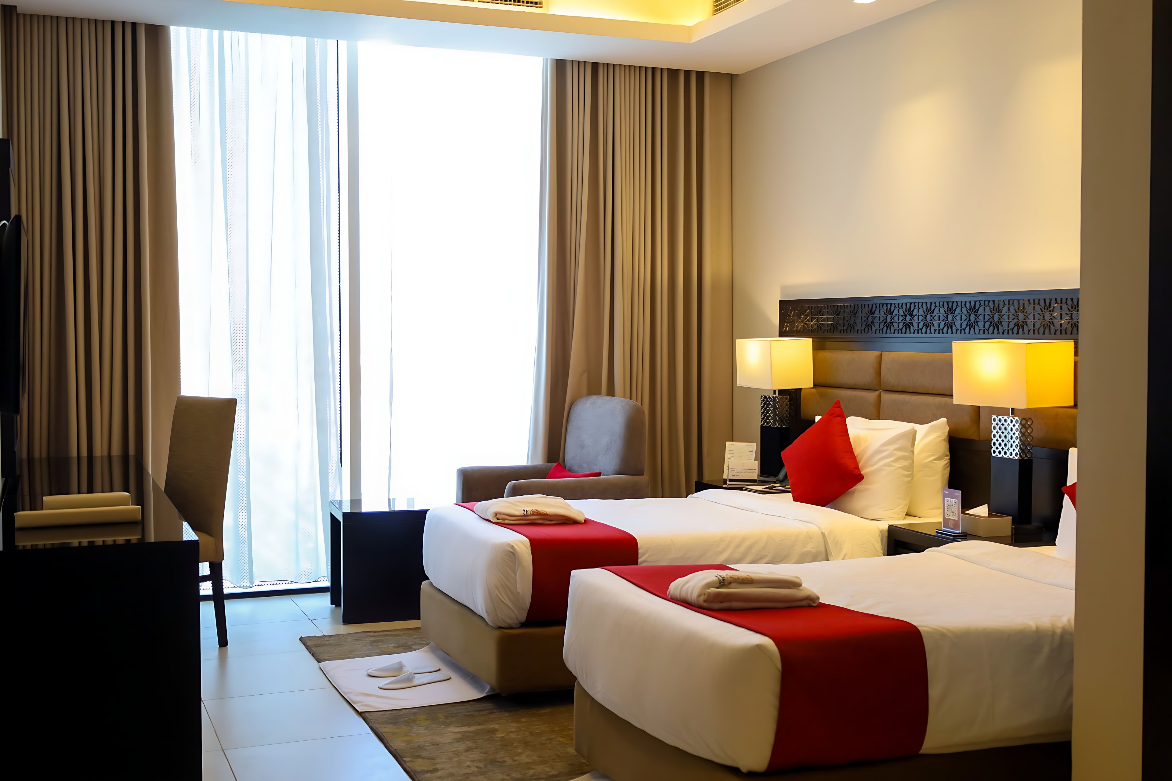 A hotel room with two beds, a desk, a chair, and a view of the outdoors.