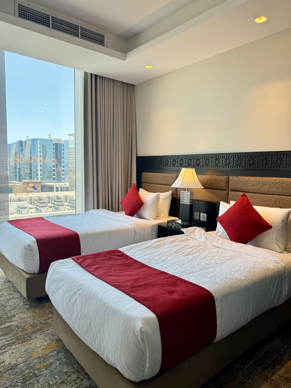 A hotel room with two beds, a large window with a city view, a headboard with a decorative panel, and a nightstand with a lamp.