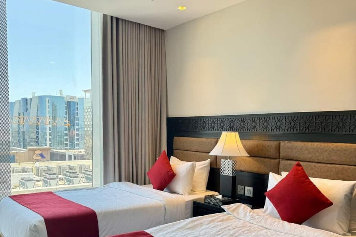 A hotel room with two beds, a large window with a city view, a headboard with a decorative panel, and a nightstand with a lamp.