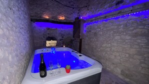 Un bain à remous avec une bouteille et des verres, placé devant un mur de pierre éclairé de lumière bleue.