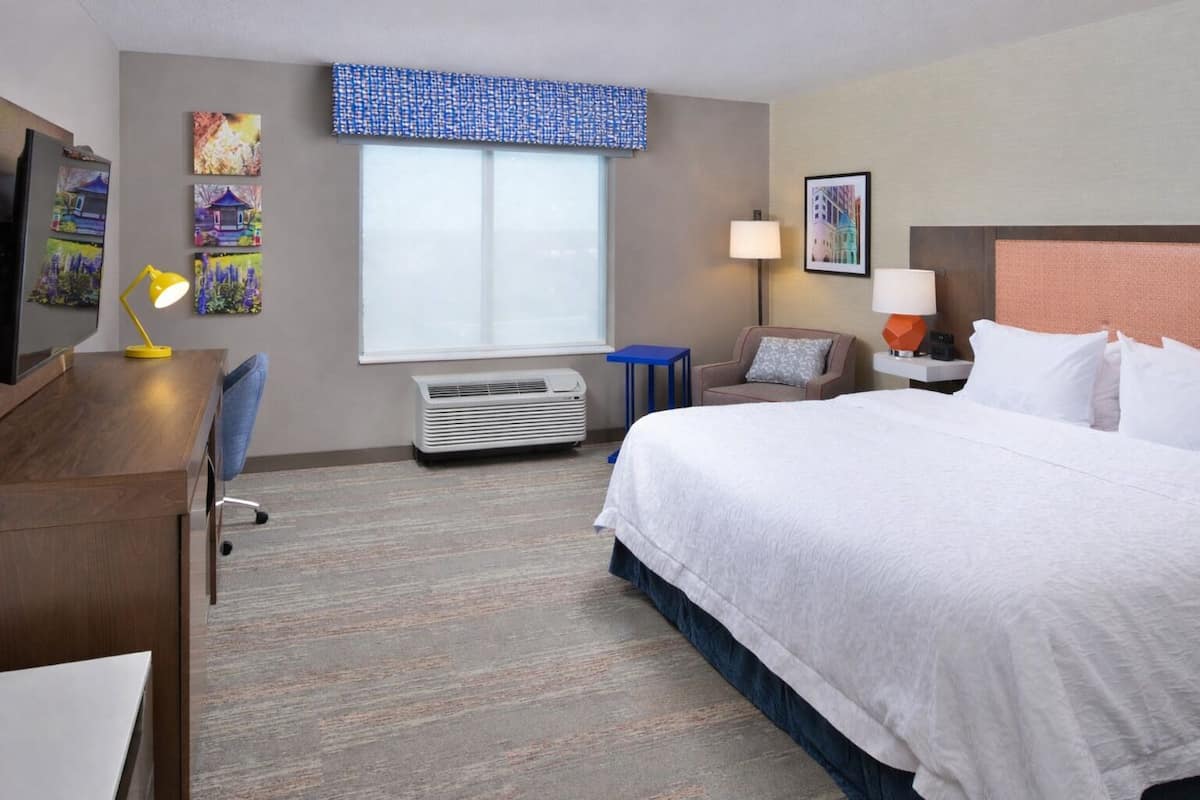 A hotel room with a bed, desk, chair, air conditioner, window, and artwork on the walls.