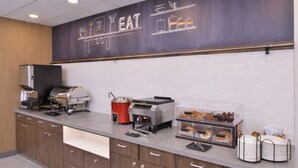 Uma cozinha compacta com um quadro negro com a inscrição 'EAT', uma churrasqueira, uma máquina de café e uma vitrine com doces.