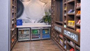 Um corredor estreito com prateleiras de madeira em ambos os lados, um balcão central com uma planta e três vitrines refrigeradas.
