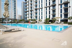 Piscina com espreguiçadeiras e guarda-sóis, cercada por um edifício moderno com varandas.
