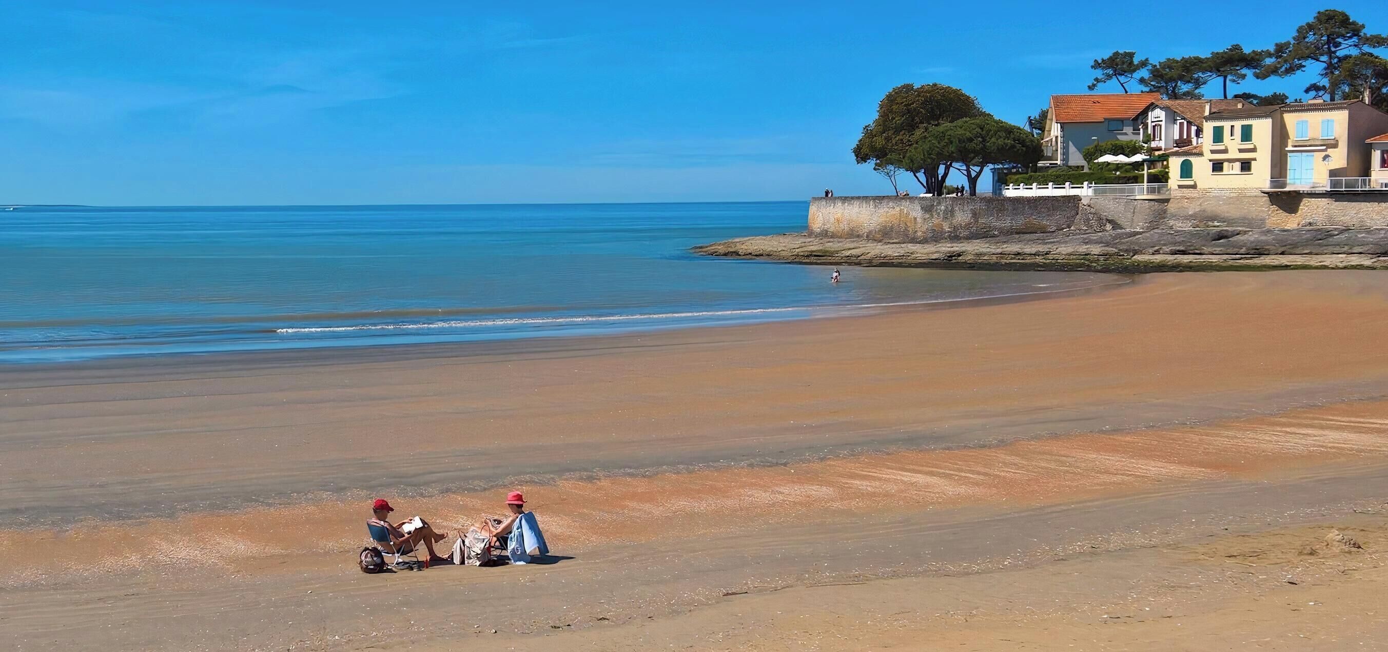 A beach with a clear sky, calm sea, and a few people relaxing. There are buildings with a traditional architectural style in the background.