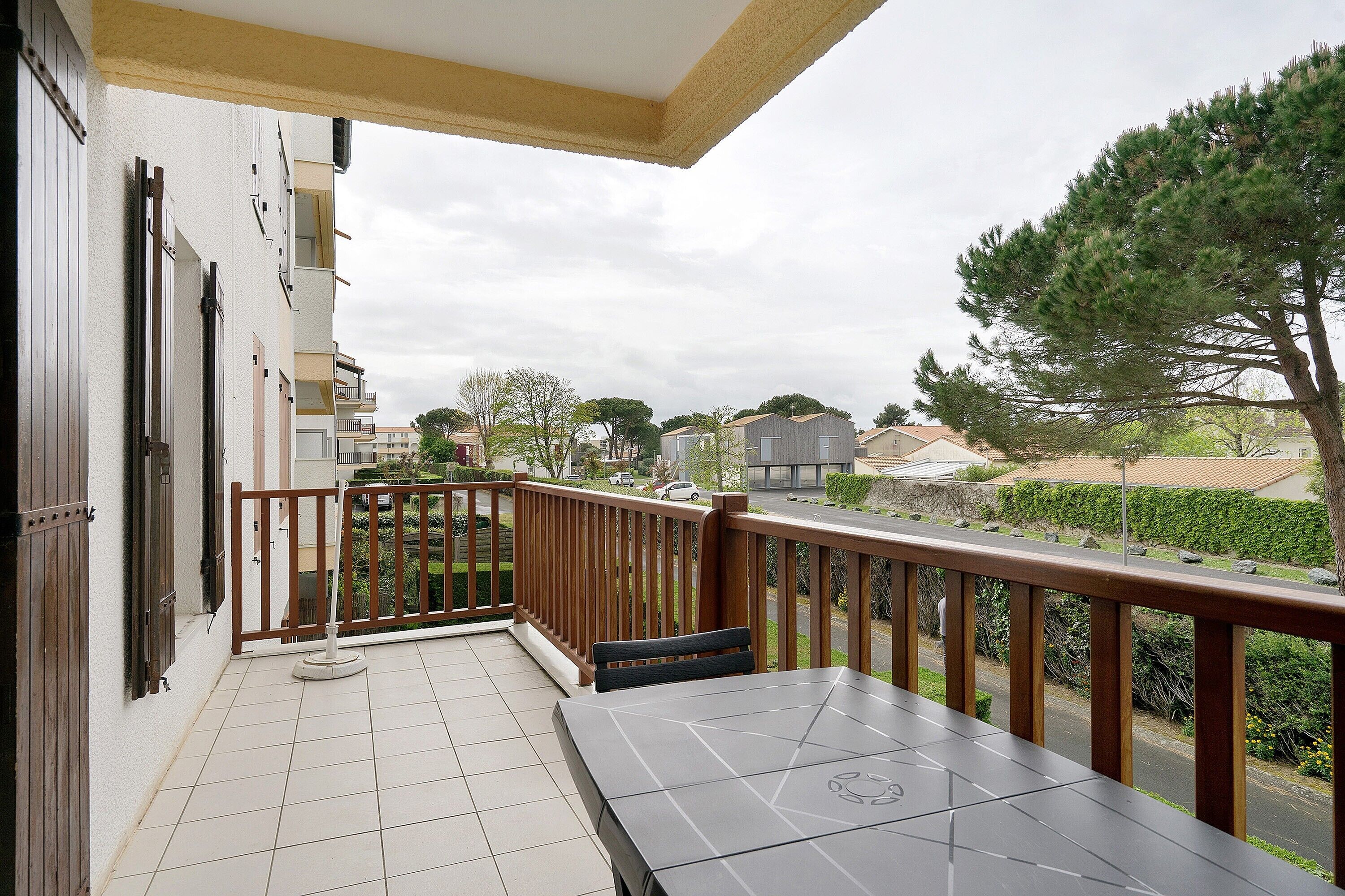 Un balcon avec une table et des chaises, une balustrade en bois et une vue sur un quartier résidentiel avec des maisons et des arbres.