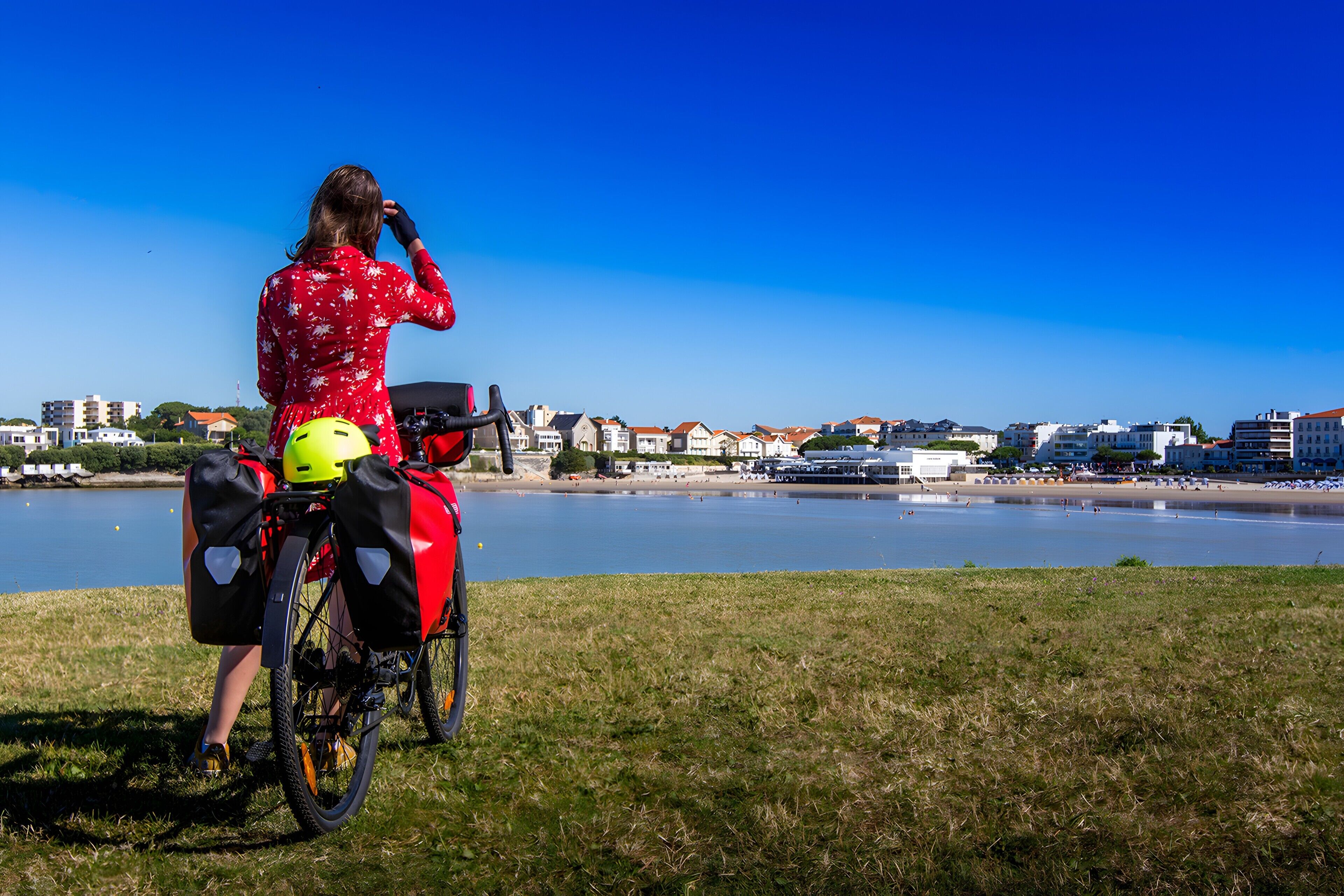 Une personne à vélo, portant un casque jaune et des sacoches, surplombe une étendue d’eau avec des bâtiments en arrière-plan.