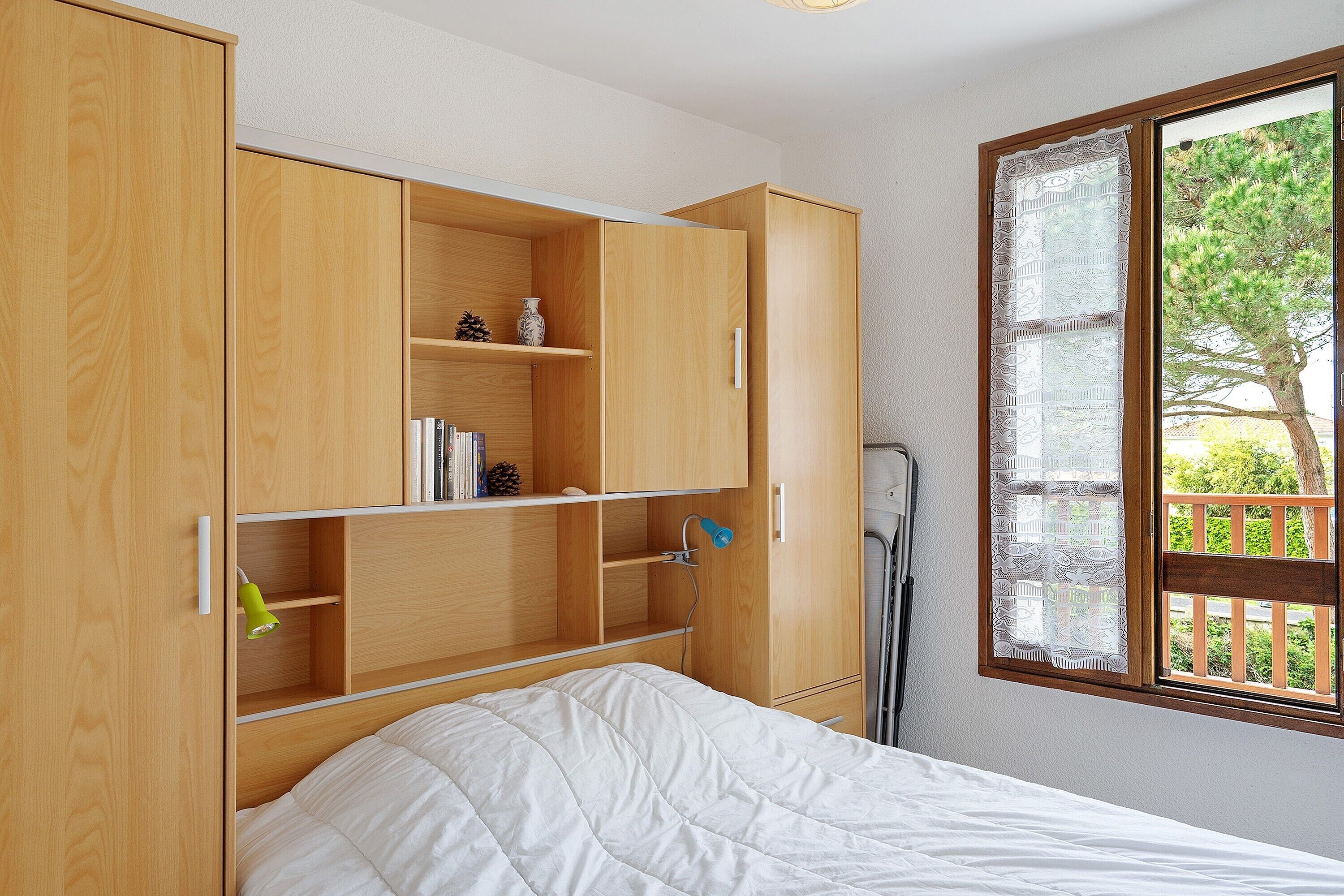 A bedroom with a bed, wooden wardrobe, and a window with a view of greenery.