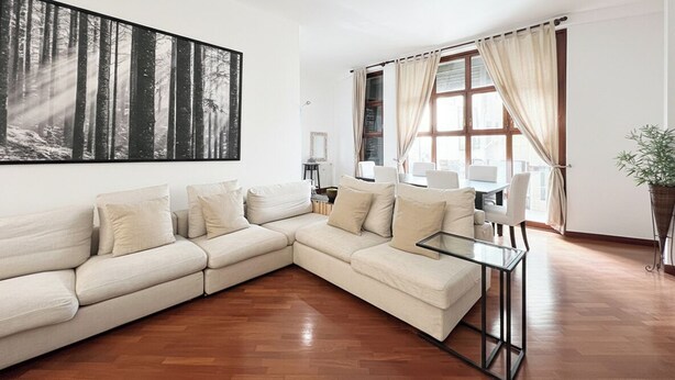 Ein modernes Wohnzimmer mit einem Ecksofa, einem Glastisch, einem großen Wandbild mit Waldmotiv und einem Essbereich mit Tisch und Stühlen.