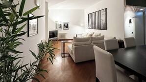Ein modernes Wohnzimmer mit einer Sofagarnitur, einem Esstisch und einem Fernseher. An den Wänden befinden sich gerahmte Bilder und in der Ecke steht eine Pflanze.