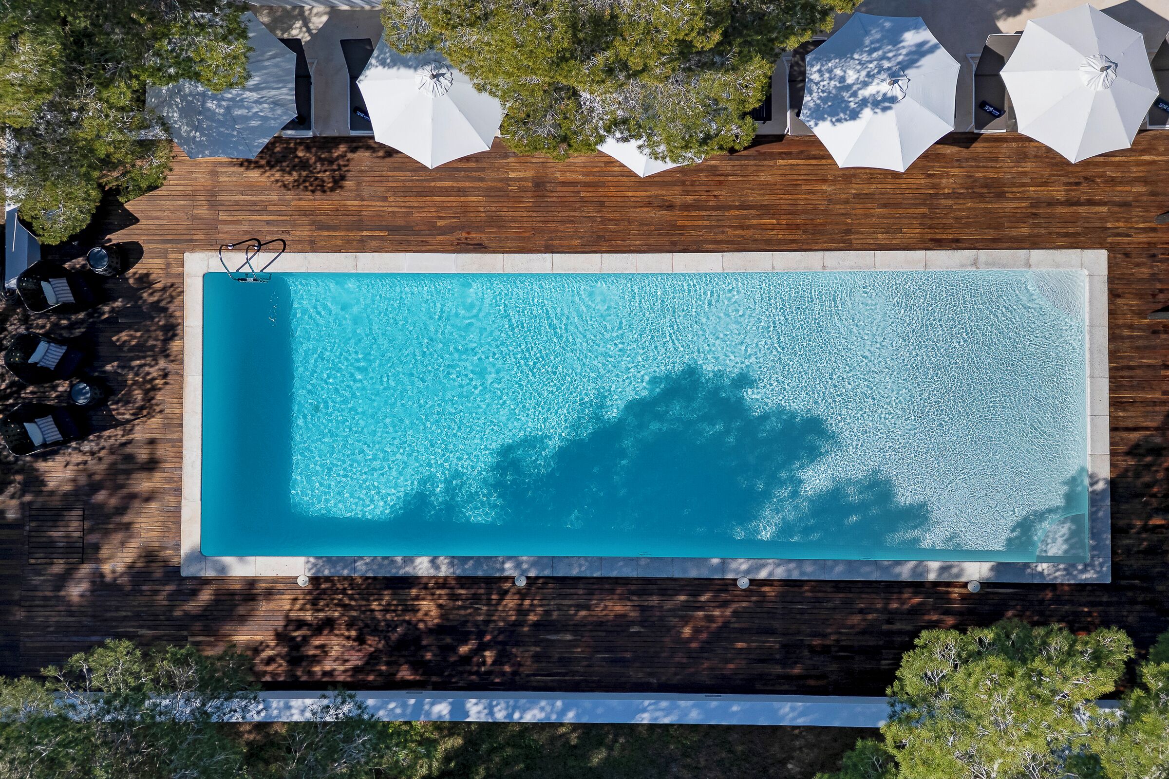 Une vue aérienne d’une piscine rectangulaire entourée d’une terrasse en bois, agrémentée de parasols blancs et de chaises longues.