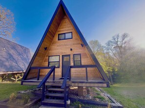 Una cabaña de madera triangular con un porche y escaleras, rodeada de árboles.