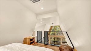 A bedroom with a large bed, a wooden nightstand, a brown leather pouf, and a view of a balcony with a wooden railing. - White Oak Haven-Cozy Forest A-frame near Bentonville (Benton County)